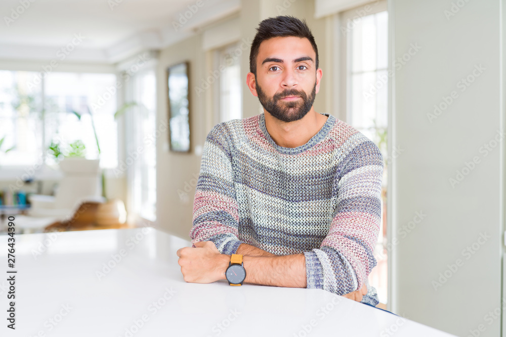 Handsome hispanic man wearing casual sweater at home with serious expression on face. Simple and natural looking at the camera. Handsome hispanic man wearing casual sweater at home with serious expression on face. Simple and natural looking at the camera.