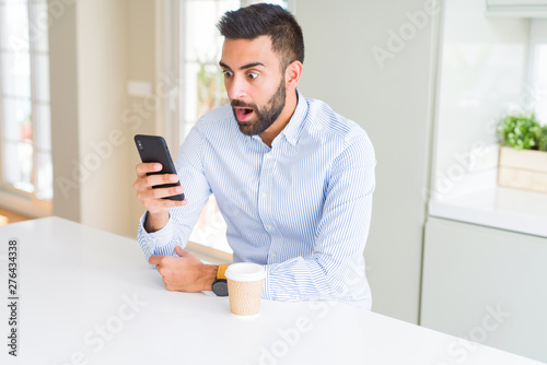 Handsome hispanic business man drinking coffee and using smartphone scared in shock with a surprise face, afraid and excited with fear expression