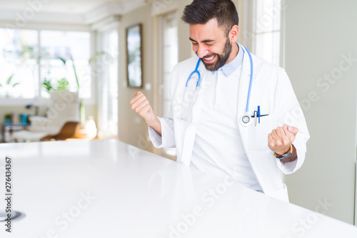 Handsome hispanic doctor man wearing stethoscope at the clinic very happy and excited doing winner gesture with arms raised  smiling and screaming for success. Celebration concept.
