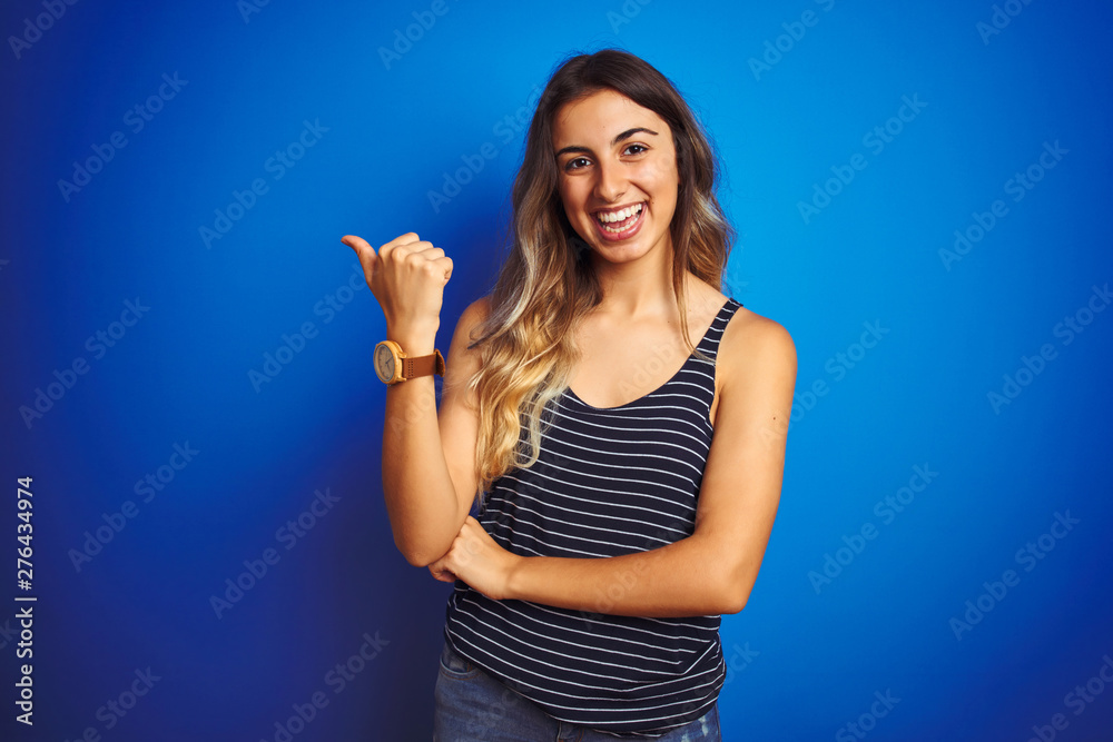 Fototapeta premium Young beautiful woman wearing stripes t-shirt over blue isolated background smiling with happy face looking and pointing to the side with thumb up.