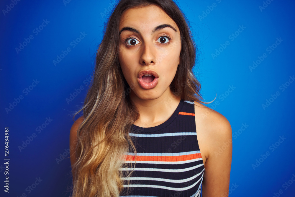 Young beautiful woman wearing stripes t-shirt over blue isolated background scared in shock with a surprise face, afraid and excited with fear expression Young beautiful woman wearing stripes t-shirt over blue isolated background scared in shock with a surprise face, afraid and excited with fear expression