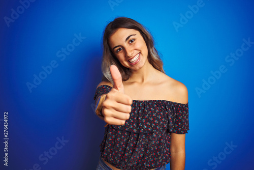 Young beautiful woman wearing floral t-shirt over blue isolated background doing happy thumbs up gesture with hand. Approving expression looking at the camera with showing success.