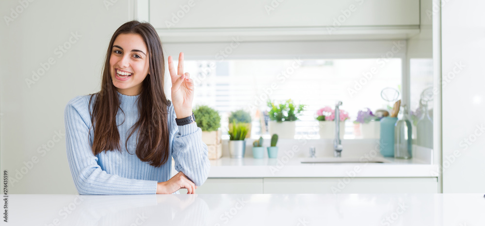 Wide angle picture of beautiful young woman sitting on white table at home smiling with happy face winking at the camera doing victory sign. Number two. Wide angle picture of beautiful young woman sitting on white table at home smiling with happy face winking at the camera doing victory sign. Number two.