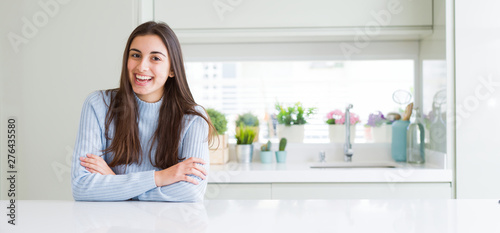 Wide angle picture of beautiful young woman sitting on white table at home happy face smiling with crossed arms looking at the camera. Positive person.