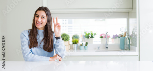 Wide angle picture of beautiful young woman sitting on white table at home smiling with happy face winking at the camera doing victory sign. Number two.