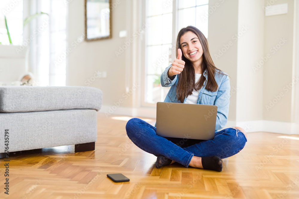 Beautiful young woman sitting on the floor with crossed legs using laptop happy with big smile doing ok sign, thumb up with fingers, excellent sign Beautiful young woman sitting on the floor with crossed legs using laptop happy with big smile doing ok sign, thumb up with fingers, excellent sign
