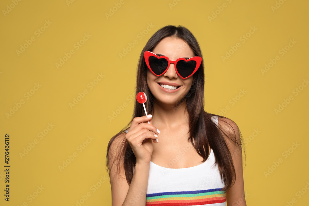 Fototapeta premium Portrait of beautiful young woman with heart shaped sunglasses and lollipop on color background