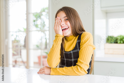 Young beautiful blonde kid girl wearing casual yellow sweater at home covering one eye with hand, confident smile on face and surprise emotion.