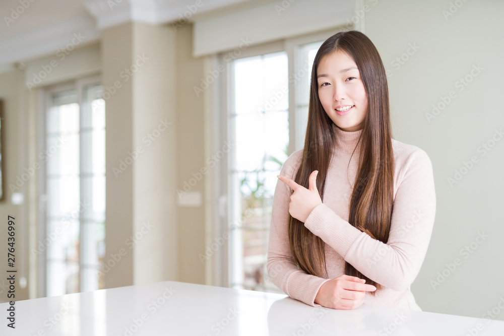 Beautiful Asian woman wearing casual sweater on white table cheerful with a smile of face pointing with hand and finger up to the side with happy and natural expression on face Beautiful Asian woman wearing casual sweater on white table cheerful with a smile of face pointing with hand and finger up to the side with happy and natural expression on face
