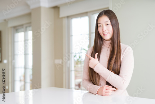 Beautiful Asian woman wearing casual sweater on white table cheerful with a smile of face pointing with hand and finger up to the side with happy and natural expression on face