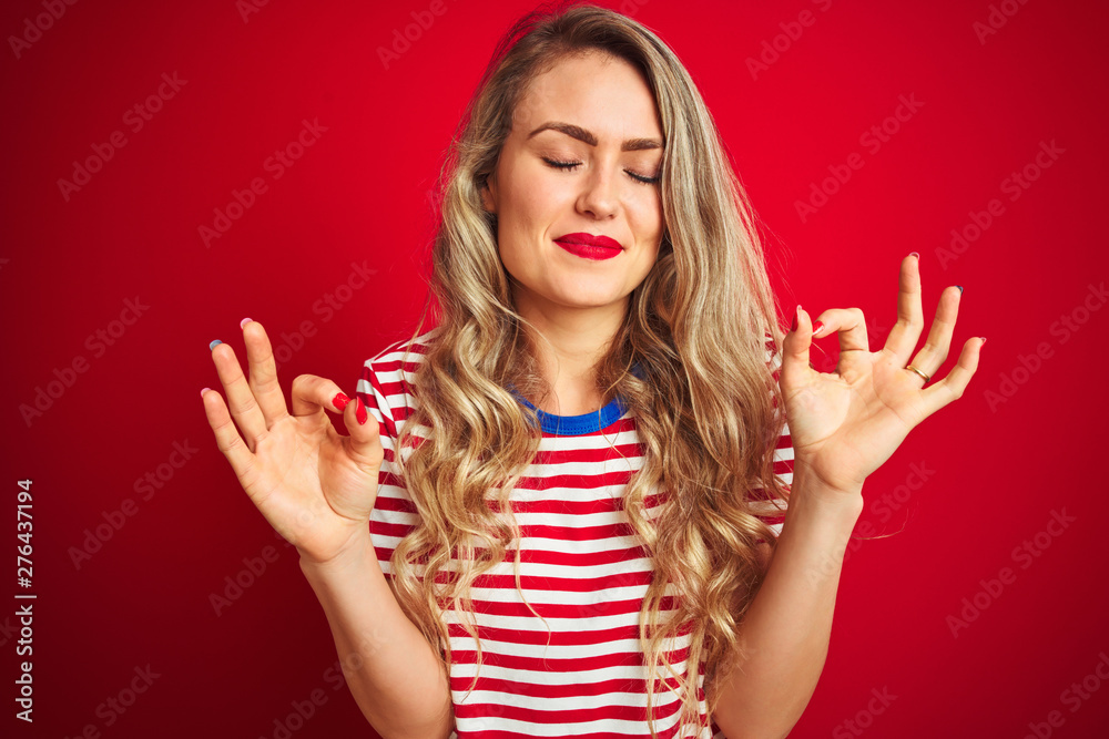 Young beautiful woman wearing stripes t-shirt standing over red isolated background relax and smiling with eyes closed doing meditation gesture with fingers. Yoga concept. Young beautiful woman wearing stripes t-shirt standing over red isolated background relax and smiling with eyes closed doing meditation gesture with fingers. Yoga concept.