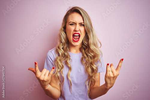 Young beautiful woman wearing purple t-shirt standing over pink isolated background shouting with crazy expression doing rock symbol with hands up. Music star. Heavy concept.