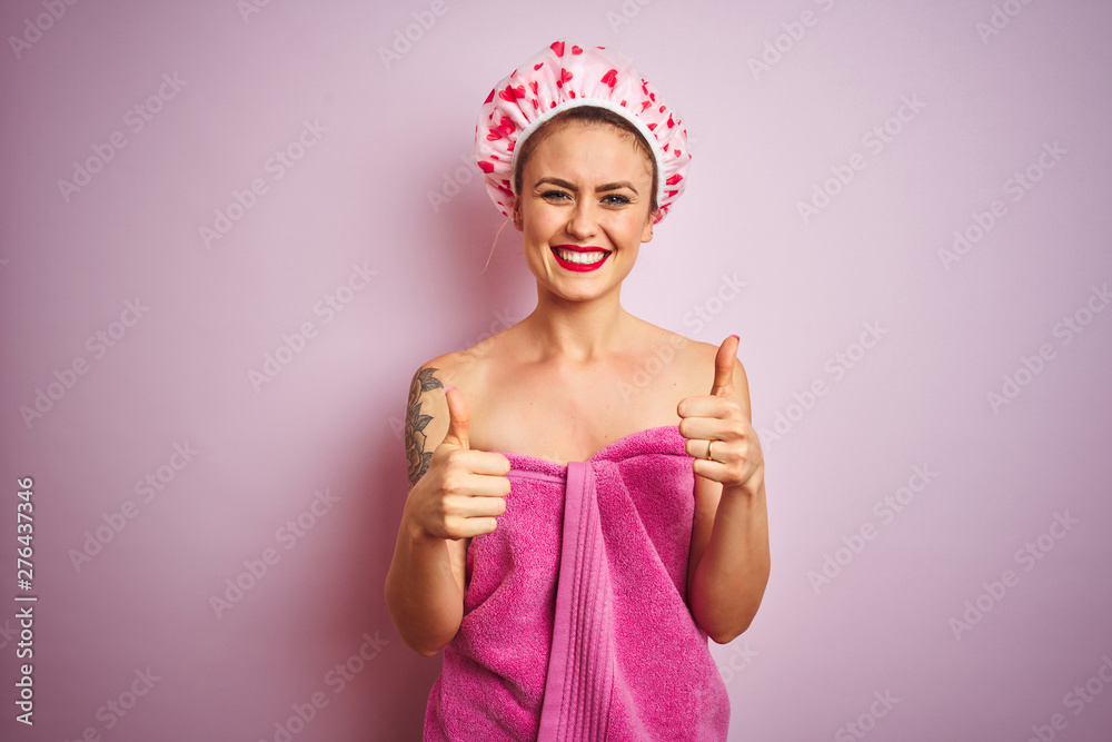 Young beautiful woman wearing towel and bath hat after shower over pink isolated background success sign doing positive gesture with hand, thumbs up smiling and happy. Cheerful expression 