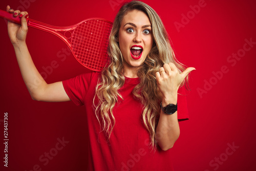 Young sportwoman playing tennis with racket and a ball over red isolated background pointing and showing with thumb up to the side with happy face smiling