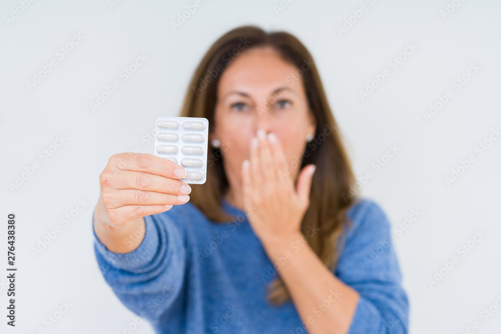 Middle age woman holding pharmaceutical pills over isolated background cover mouth with hand shocked with shame for mistake, expression of fear, scared in silence, secret concept Middle age woman holding pharmaceutical pills over isolated background cover mouth with hand shocked with shame for mistake, expression of fear, scared in silence, secret concept