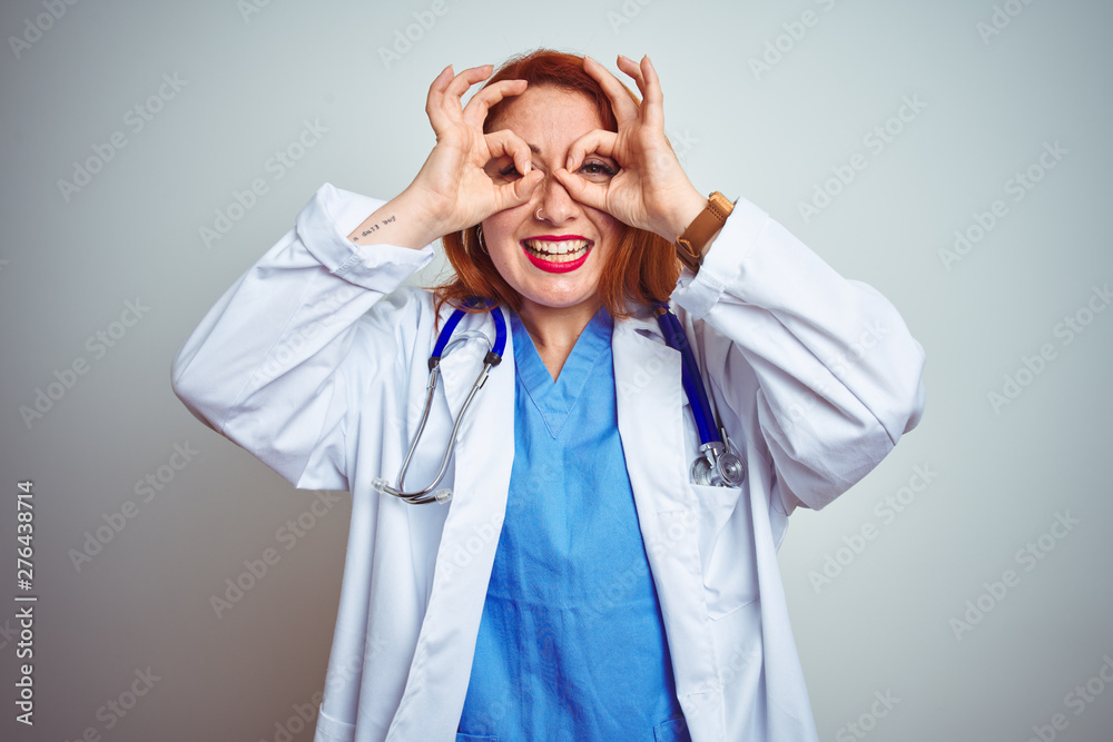 Young redhead doctor woman using stethoscope over white isolated background doing ok gesture like binoculars sticking tongue out, eyes looking through fingers. Crazy expression. Young redhead doctor woman using stethoscope over white isolated background doing ok gesture like binoculars sticking tongue out, eyes looking through fingers. Crazy expression.