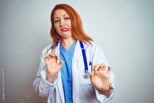 Young redhead doctor woman using stethoscope over white isolated background disgusted expression  displeased and fearful doing disgust face because aversion reaction. With hands raised.