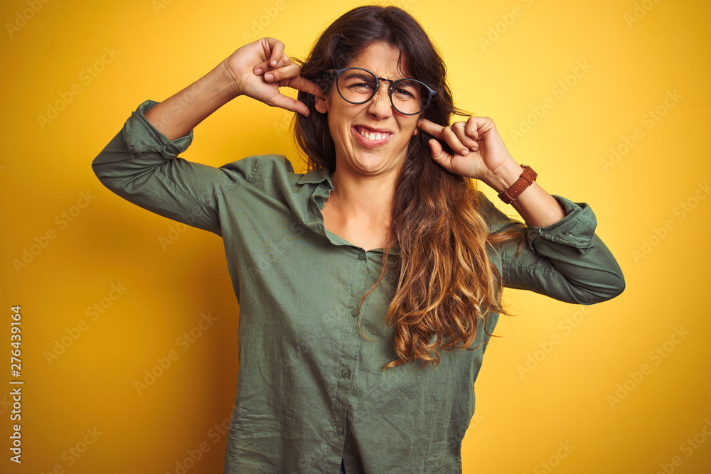 Young beautiful woman wearing green shirt and glasses over yelllow isolated background covering ears with fingers with annoyed expression for the noise of loud music. Deaf concept. Young beautiful woman wearing green shirt and glasses over yelllow isolated background covering ears with fingers with annoyed expression for the noise of loud music. Deaf concept.