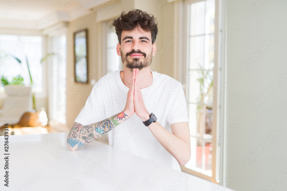 Young man wearing casual t-shirt sitting on white table praying with hands together asking for forgiveness smiling confident.