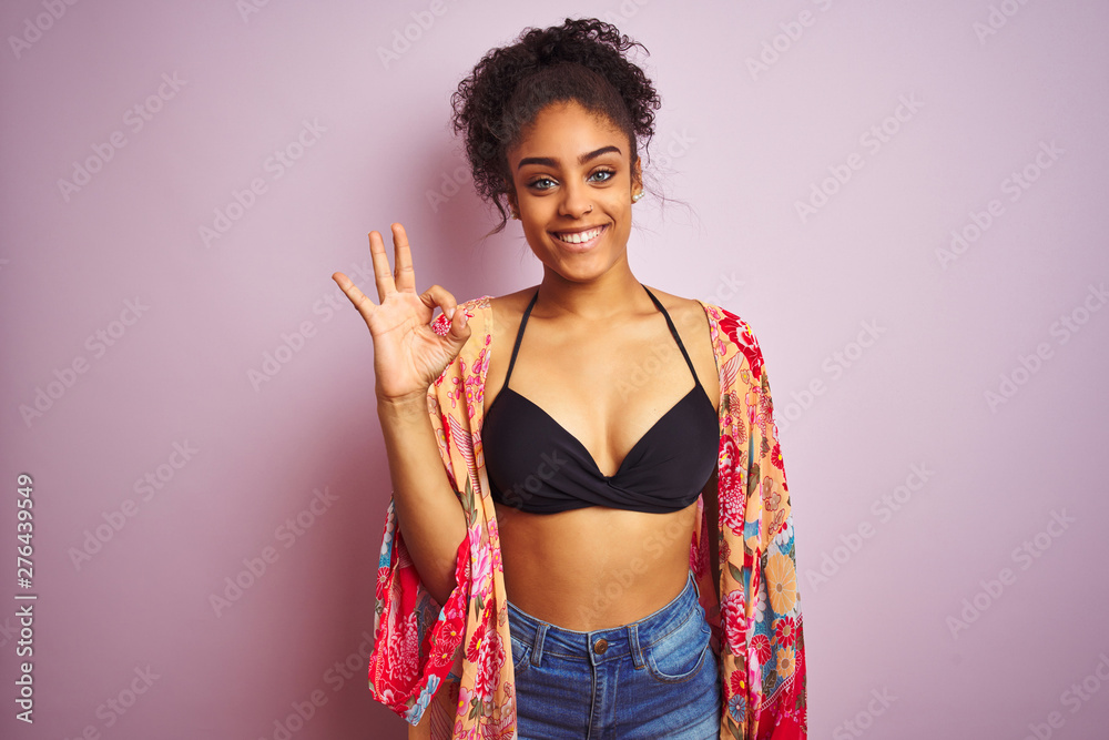 American woman on holiday wearing bikini and colorful dress over isolated pink background smiling positive doing ok sign with hand and fingers. Successful expression. American woman on holiday wearing bikini and colorful dress over isolated pink background smiling positive doing ok sign with hand and fingers. Successful expression.