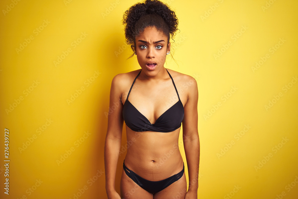 African american woman on vacation wearing bikini standing over isolated yellow background afraid and shocked with surprise and amazed expression, fear and excited face. African american woman on vacation wearing bikini standing over isolated yellow background afraid and shocked with surprise and amazed expression, fear and excited face.