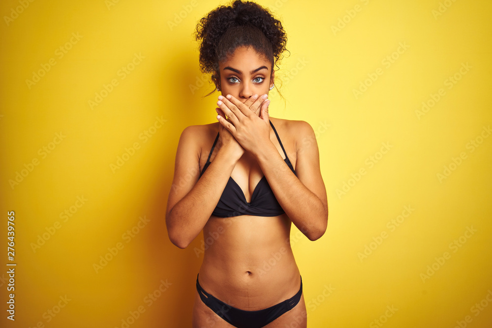 African american woman on vacation wearing bikini standing over isolated yellow background shocked covering mouth with hands for mistake. Secret concept. - obrazy, fototapety, plakaty 