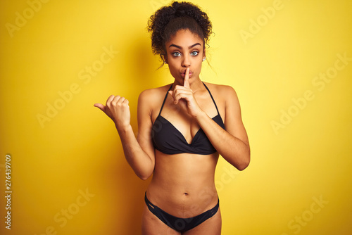 African american woman on vacation wearing bikini standing over isolated yellow background asking to be quiet with finger on lips pointing with hand to the side. Silence and secret concept.