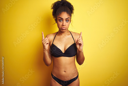 African american woman on vacation wearing bikini standing over isolated yellow background Pointing up looking sad and upset, indicating direction with fingers, unhappy and depressed. © Krakenimages.com
