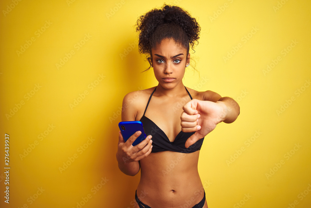 African american woman wearing bikini using smartphone over isolated yellow background with angry face, negative sign showing dislike with thumbs down, rejection concept African american woman wearing bikini using smartphone over isolated yellow background with angry face, negative sign showing dislike with thumbs down, rejection concept