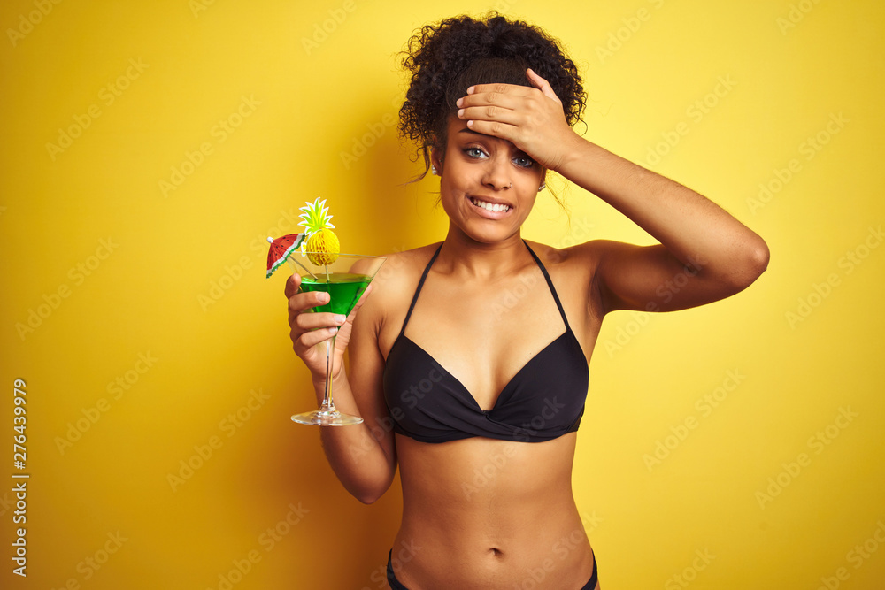 Afro woman on vacation wearing bikini drinking a cocktail over isolated yellow background stressed with hand on head, shocked with shame and surprise face, angry and frustrated. Afro woman on vacation wearing bikini drinking a cocktail over isolated yellow background stressed with hand on head, shocked with shame and surprise face, angry and frustrated.