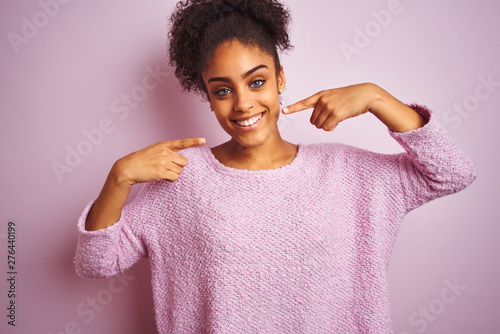 Young african american woman wearing winter sweater standing over isolated pink background smiling cheerful showing and pointing with fingers teeth and mouth. Dental health concept.