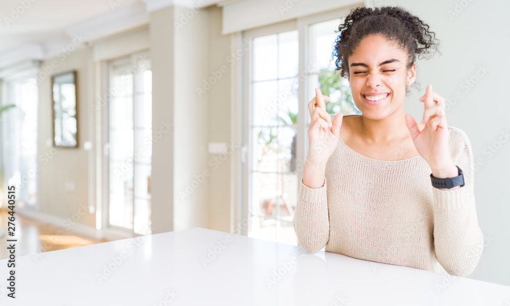 Beautiful young african american woman with afro hair sitting on table at home smiling crossing fingers with hope and eyes closed. Luck and superstitious concept. Beautiful young african american woman with afro hair sitting on table at home smiling crossing fingers with hope and eyes closed. Luck and superstitious concept.