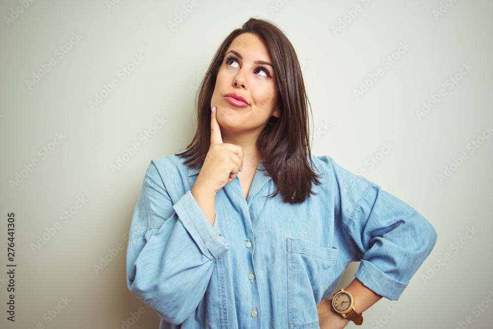 Young beautiful brunette woman wearing casual blue denim shirt over isolated background with hand on chin thinking about question, pensive expression. Smiling with thoughtful face. Doubt concept. Young beautiful brunette woman wearing casual blue denim shirt over isolated background with hand on chin thinking about question, pensive expression. Smiling with thoughtful face. Doubt concept.