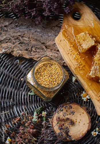 A table with a wicker table top, honey in honeycombs on a wooden board, honey in an open glass jar, with medicinal herbs oregano, chamomile and strawberry flowers. flower pollen collected by bees