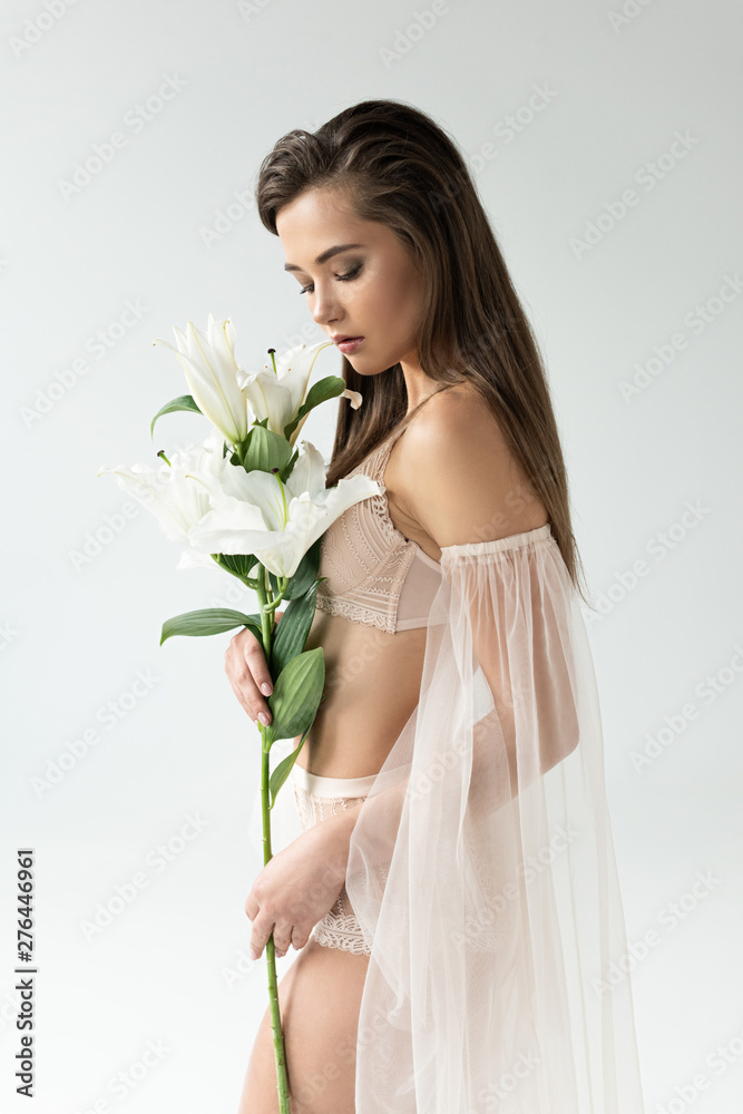 tender young woman in beige lingerie and mesh sleeves looking at lilies isolated on white tender young woman in beige lingerie and mesh sleeves looking at lilies isolated on white