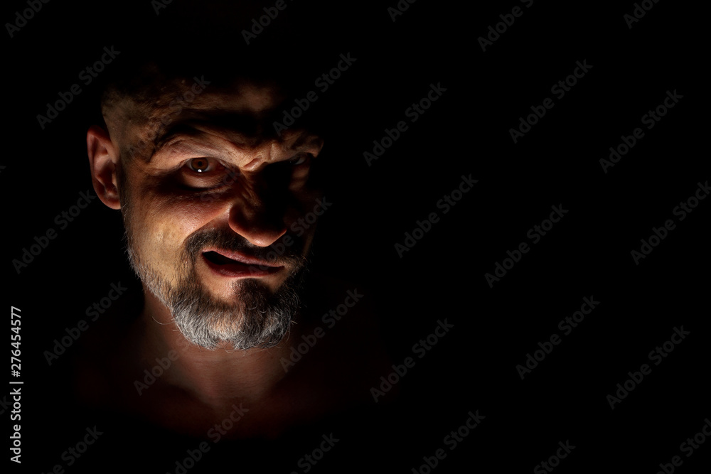 Fototapeta premium Face with a bearded man grimace against a dark background with sharp shadows. Comedic, fabulous villain or negative character conception with copy space.
