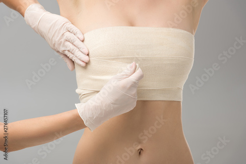 partial view of plastic surgeon in latex gloves and patient in breast bandage isolated on grey © LIGHTFIELD STUDIOS