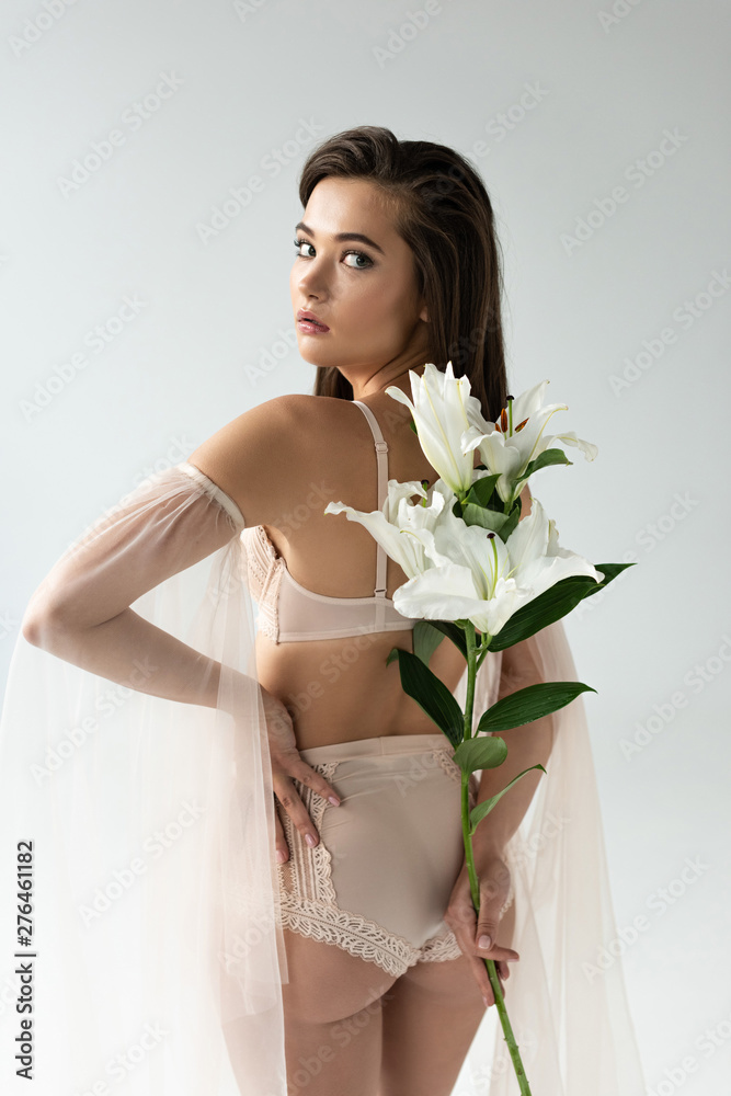 tender young woman in beige lingerie and mesh sleeves holding lilies behind back isolated on white tender young woman in beige lingerie and mesh sleeves holding lilies behind back isolated on white