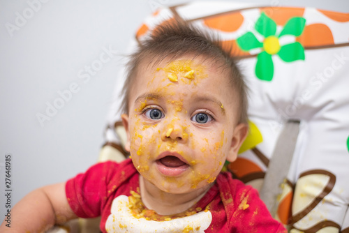 Blue-eyed baby making mess with food sitting on the highchair
