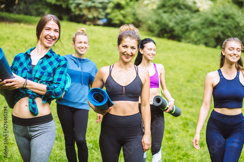 Cheerful smiling women friends in sportswear holding yoga mat and bottle in park. Middle age women going to park for fitness workout. Five curvy girls walking after exercise session outdoor at sunset.