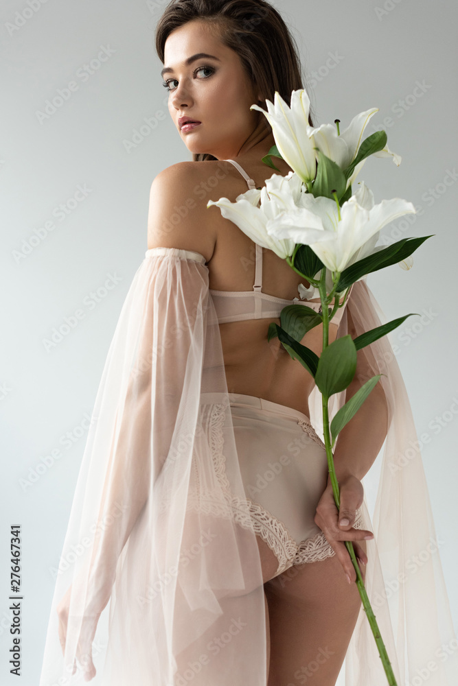 low angle view of delicate young woman in beige lingerie and mesh sleeves holding bouquet of lilies behind back isolated on white low angle view of delicate young woman in beige lingerie and mesh sleeves holding bouquet of lilies behind back isolated on white