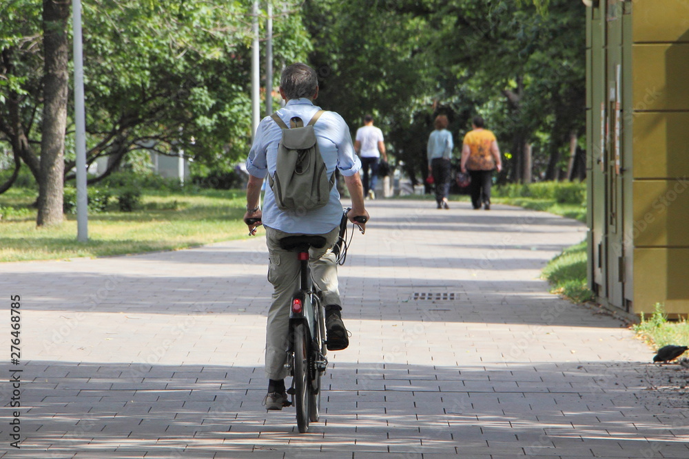 An elderly man riding a bike along the shadowy alley in the Moscow Park on a summer day, a view from back