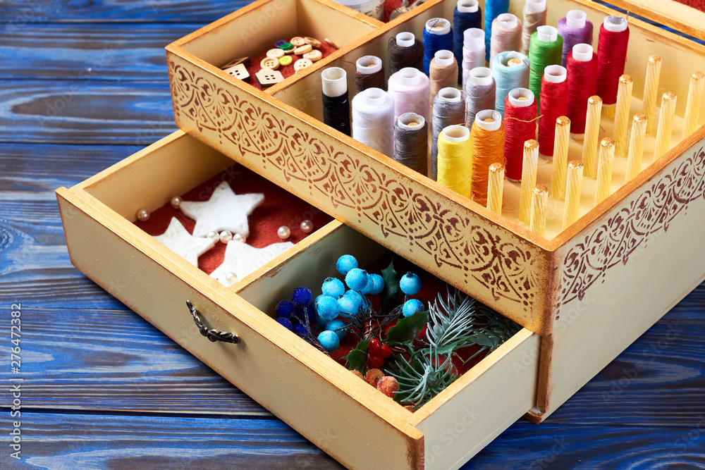 Fragment of open yellow box with pattern for sewing and needlework with spools of thread and decor and yarn balls on blue wooden background Fragment of open yellow box with pattern for sewing and needlework with spools of thread and decor and yarn balls on blue wooden background