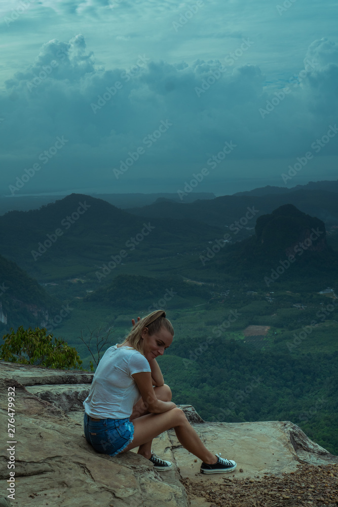 Woman taveler sitting on the cliff and looking at the beautiful landscape with mountains and valley with cloudy morning sky