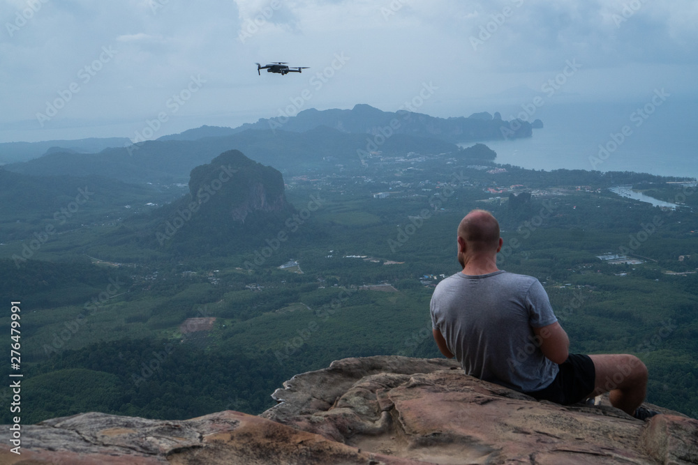 Back view of man drone operator sitting on the edge of the rock over beautiful green mountains and sky background