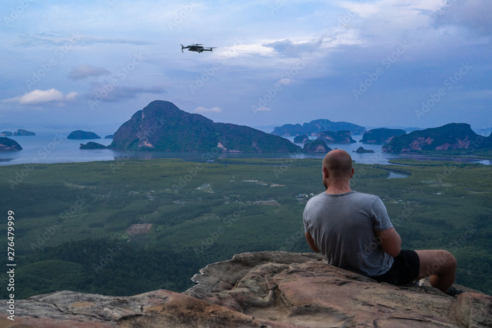 Back view of man drone operator sitting on the edge of the rock over beautiful green mountains and sky background