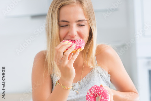 Young blonde girl eats pink donuts in home kitchen with taste emotions