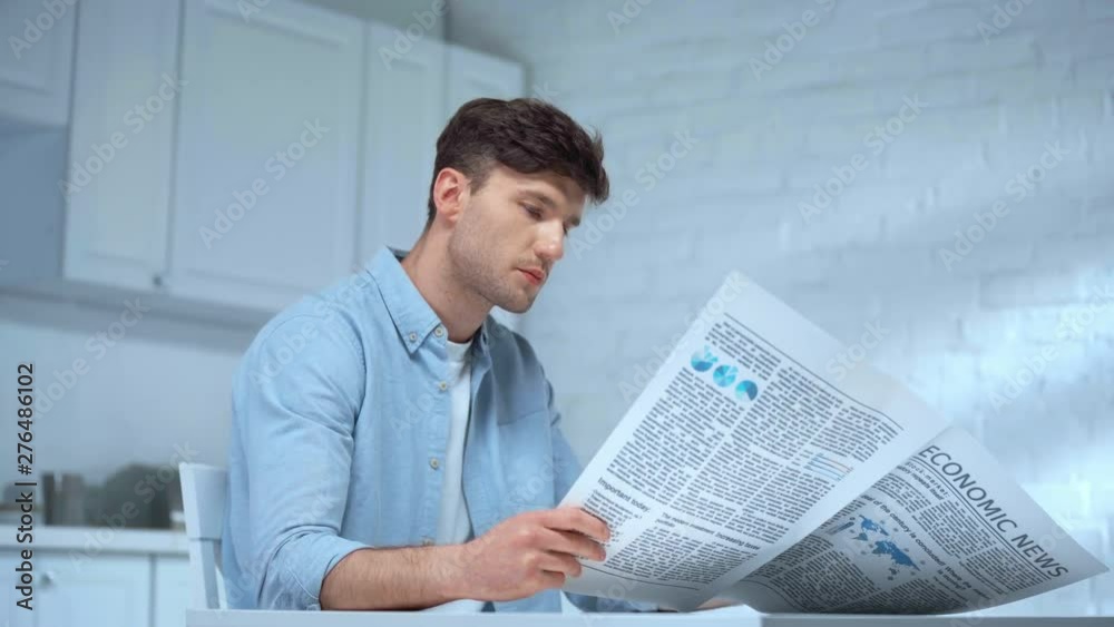 focused man in blue t-shirt drinking coffee and reading newspaper in morning in kitchen