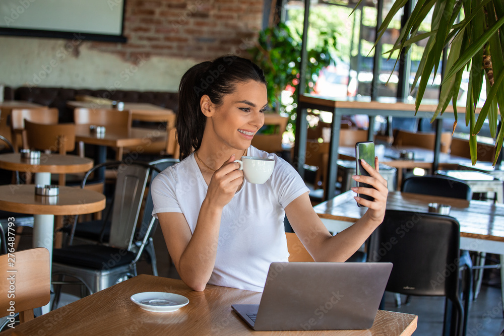 Portrait of a playful young girl taking selfie with mobile phone while sitting with laptop computer at a cafe outdoors. Morning coffee is my daily routine