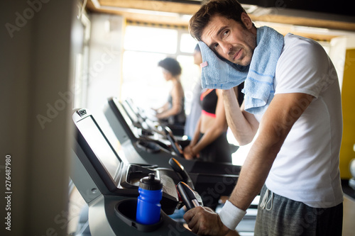 Athletic young man having rest after running training, drinking water at gym, standing at treadmill